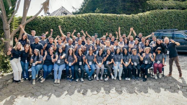 Large group photo of approximately 60 people from the Conexiam team posing together outdoors. Everyone is wearing dark gray company t-shirts with lanyards and name badges, making victory signs, peace signs, thumbs up, and enthusiastic poses in front of a tall green hedge, palm trees, and a historic building with a tiled roof.