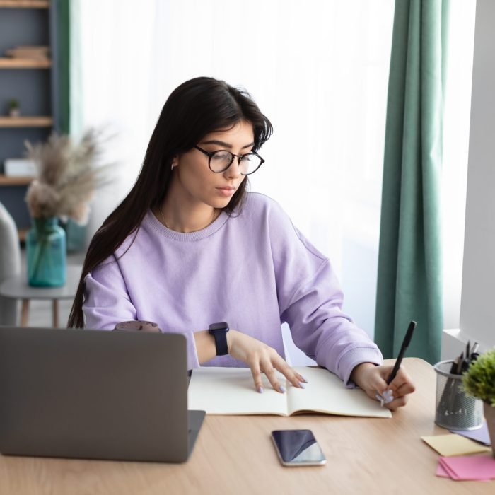 Note Taking. Portrait of focused woman in glasses sitting at table, using laptop and writing in notebook, living room interior, copy space. Female studying online from home doing remote work